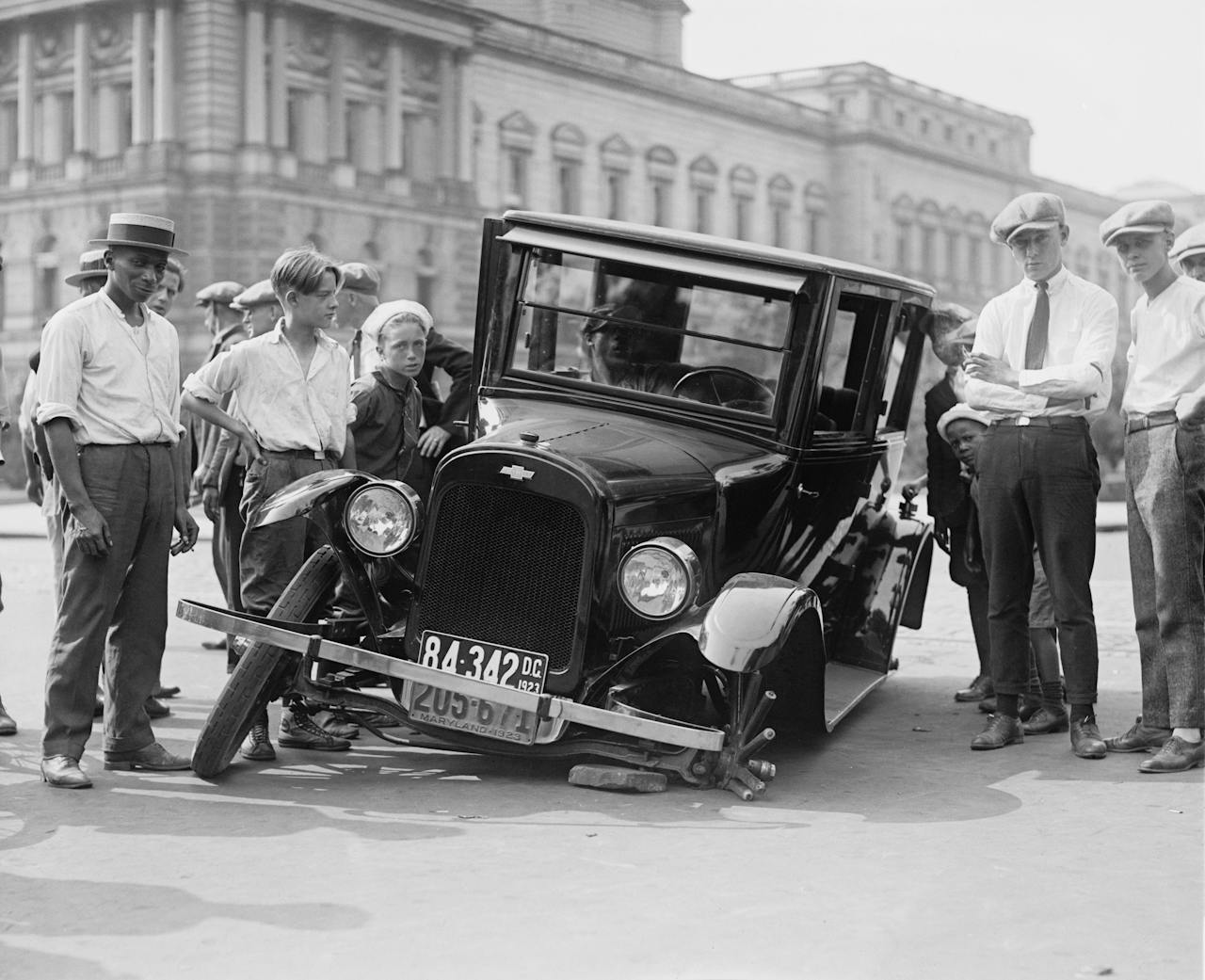 A vintage car crash attended by a group of onlookers in a 1920s urban setting.