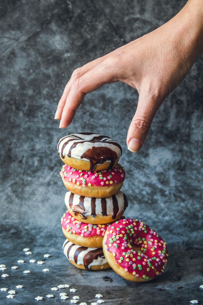 A hand reaches for a stack of colorful donuts with chocolate drizzle and sprinkles, tempting and delicious.