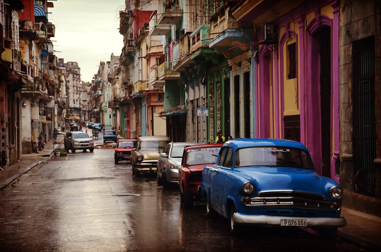 Colorful vintage cars line a wet street with vibrant buildings in a cityscape setting.